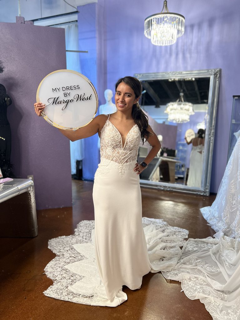 Bride in a fitted lace and crepe wedding gown holding a My Dress by Margo West sign at the Margo West Bridal Couture studio in Dallas TX